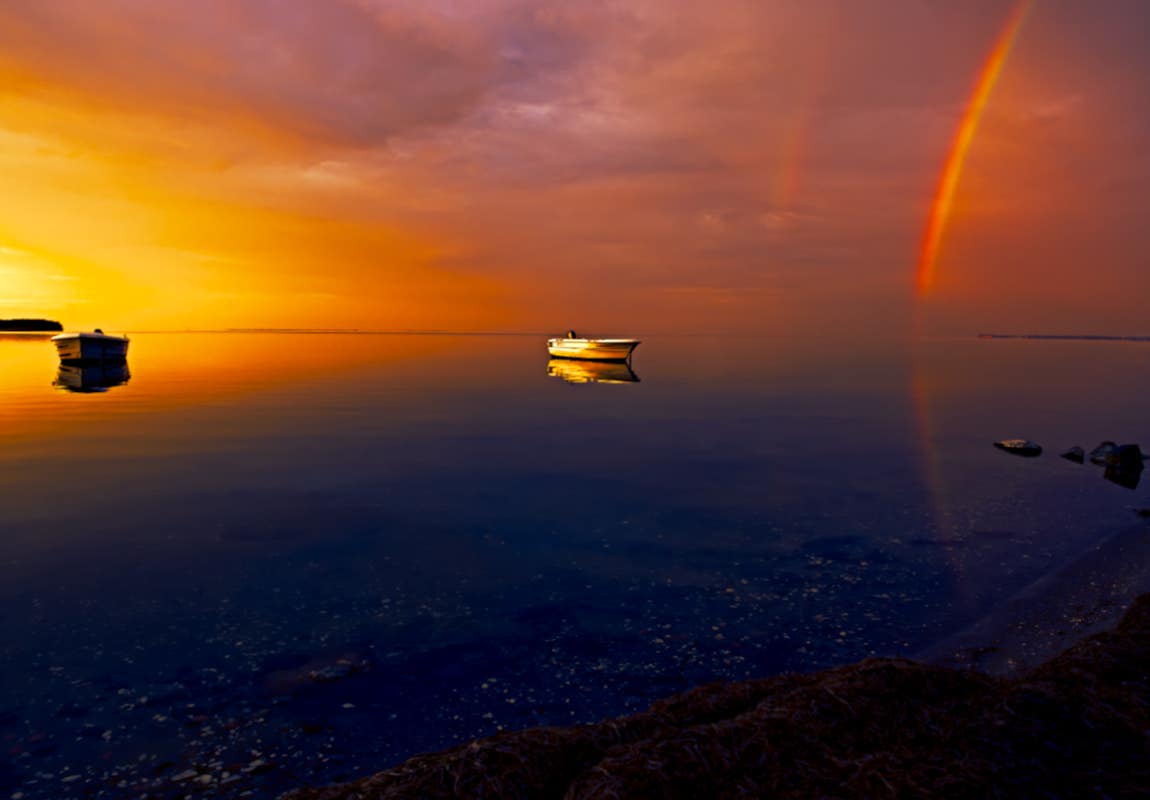 Foto eines Sonnenaufgangs mit Regenbogen und vor anker liegenden Booten