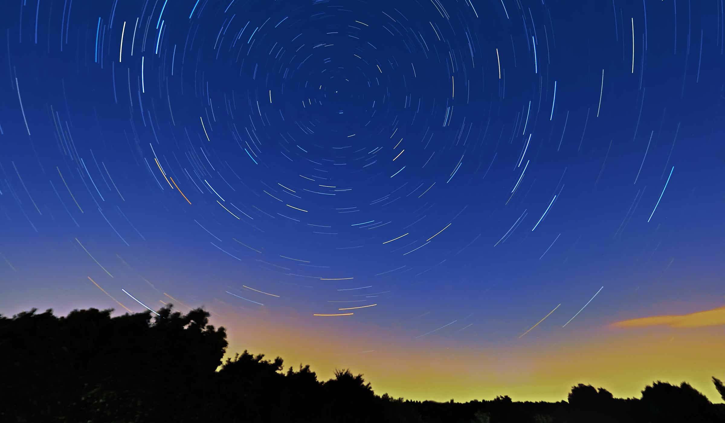 Farbaufnahme von Sternenpfaden/Star Trails mittels einer Langzeitbelichtung/Long Time Exposure von circa 30 Minuten Dauer. Der Mittelpunkt, um den sich die Sternenpfade drehen ist deutlich im Foto zu erkennen. Der Horizont befindet sich nahe des unteren Bildendes und erstrahlt in tiefem gelb/orange der in circa einer Stunde aufgehenden Sonne. Der Himmel ist wolkenlos und verläuft von tiefblau bis fast schwarz. Fotograf und Copyright: Lars König