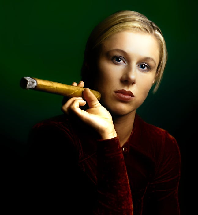 Farbaufnahme eines weiblichen Models mit einer angezündeten Zigarre in der rechten Hand. Die junge Frau ist sitzend portraitiert und stützt den rechten Ellenbogen auf dem rechten Oberschenkel ab. Sie trägt ein samtenes braunes Kleid, hat hellblonde Haare und schaut direkt in die Kamera. Der Hintergrund hat einen grünen Verlauf. Es handelt sich um eine Studioaufnahme. Fotograf und Copyright: Lars König